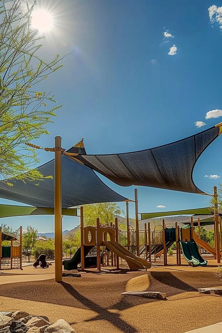 Large charcoal and green shade sails on timber-toned posts covering a desert park playground with slides and climbing structures, mountains on the horizon