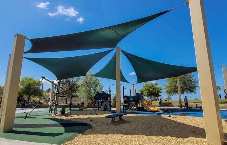 Overlapping dark green shade sails on tall steel posts spanning a community playground with climbing frames and a slide below