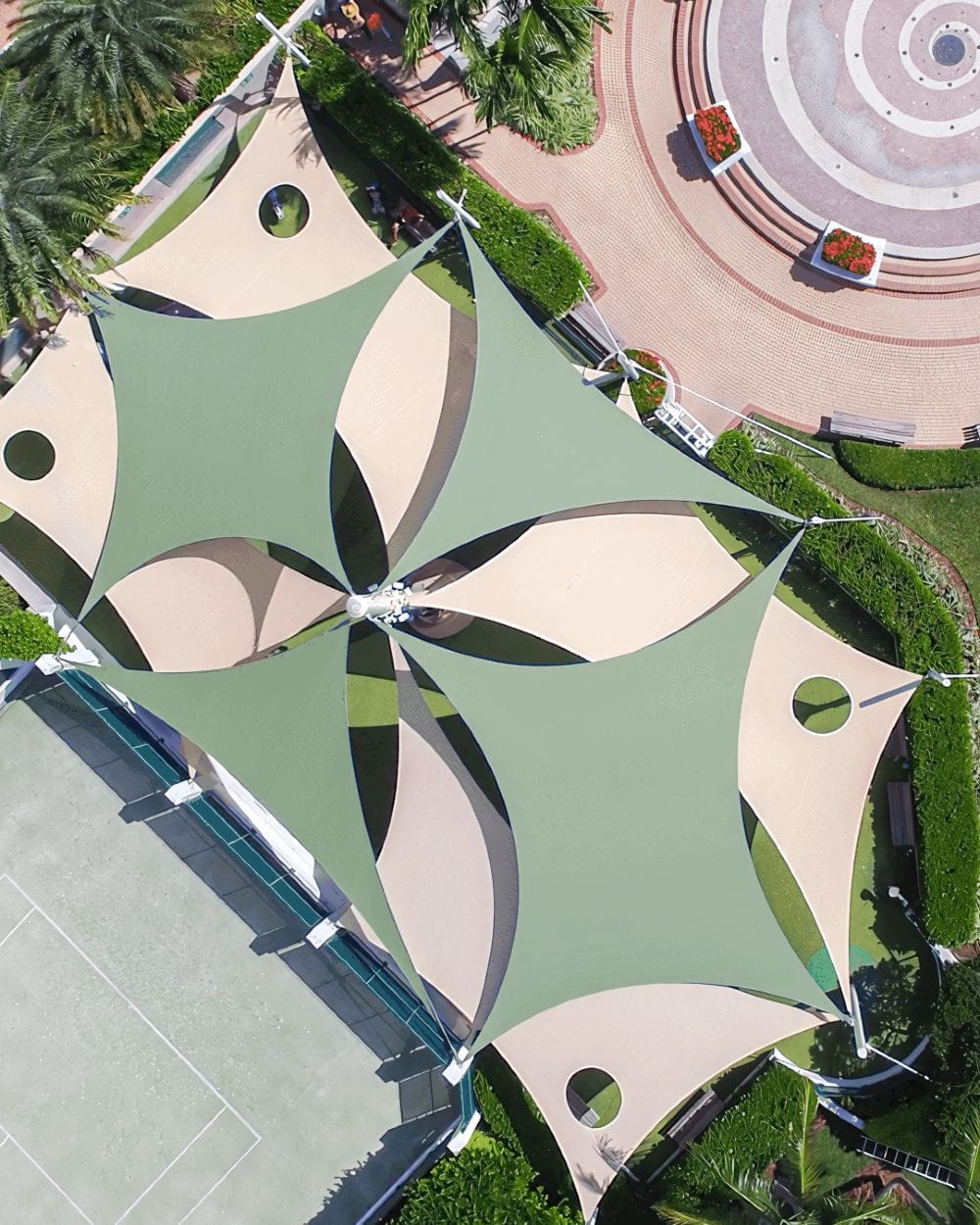 Aerial view of a cluster of sage green and sand hyperbolic paraboloid shade sails with circular vents, arranged over a resort courtyard beside a tennis court and pool