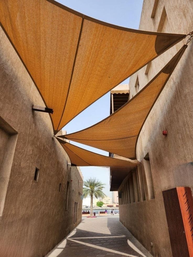 Triangular sand-coloured shade sails tensioned between the rendered walls of a heritage quarter alleyway, with a palm tree and waterfront beyond