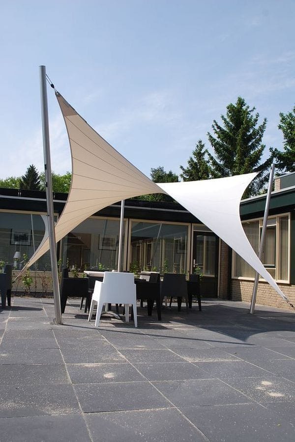 Two interlocking sand and white hyperbolic shade sails on slim stainless masts covering a dining terrace outside a low modern building
