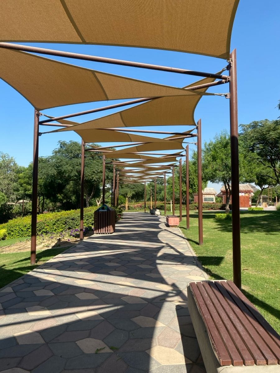 A long repeating colonnade of sand-coloured tensile membrane sails on bronze steel posts shading a paved park walkway lined with planting and benches