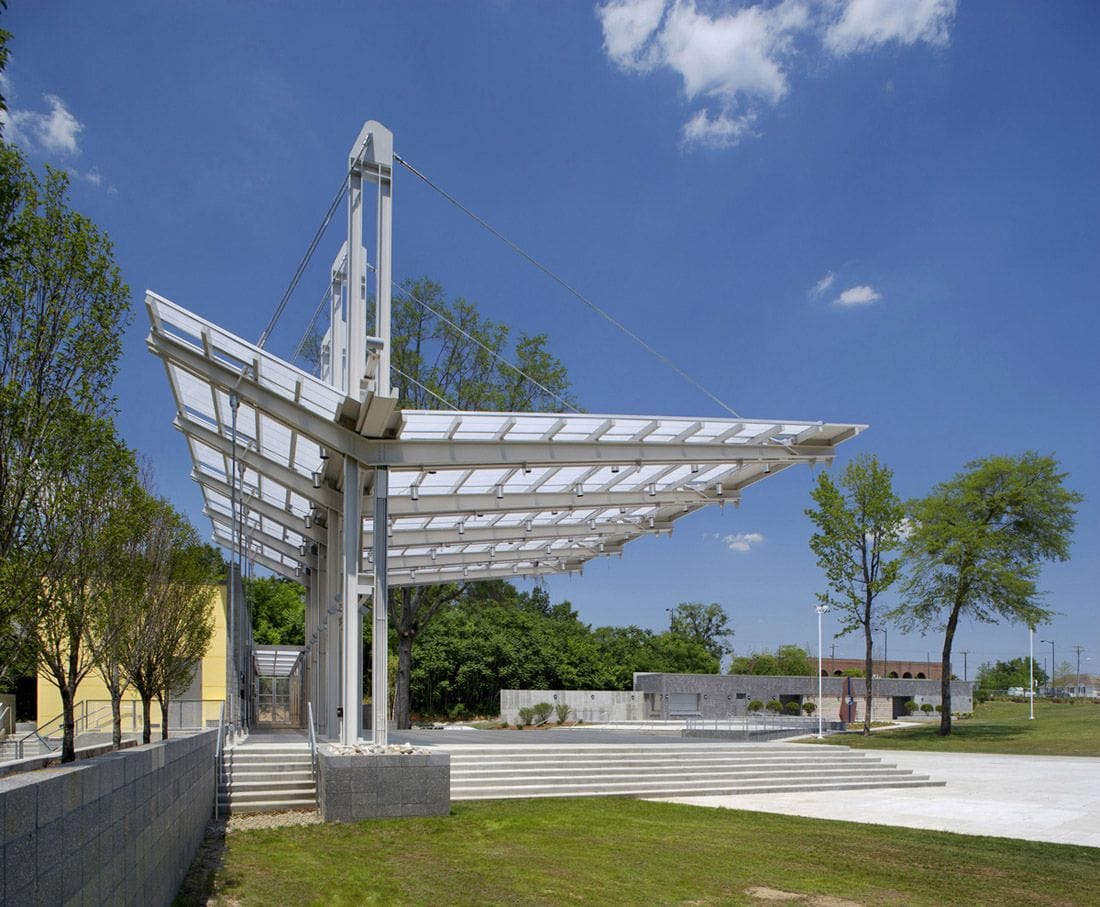 White steel cantilever canopy with translucent roof panels and tension cables projecting over a stepped public plaza against a clear blue sky